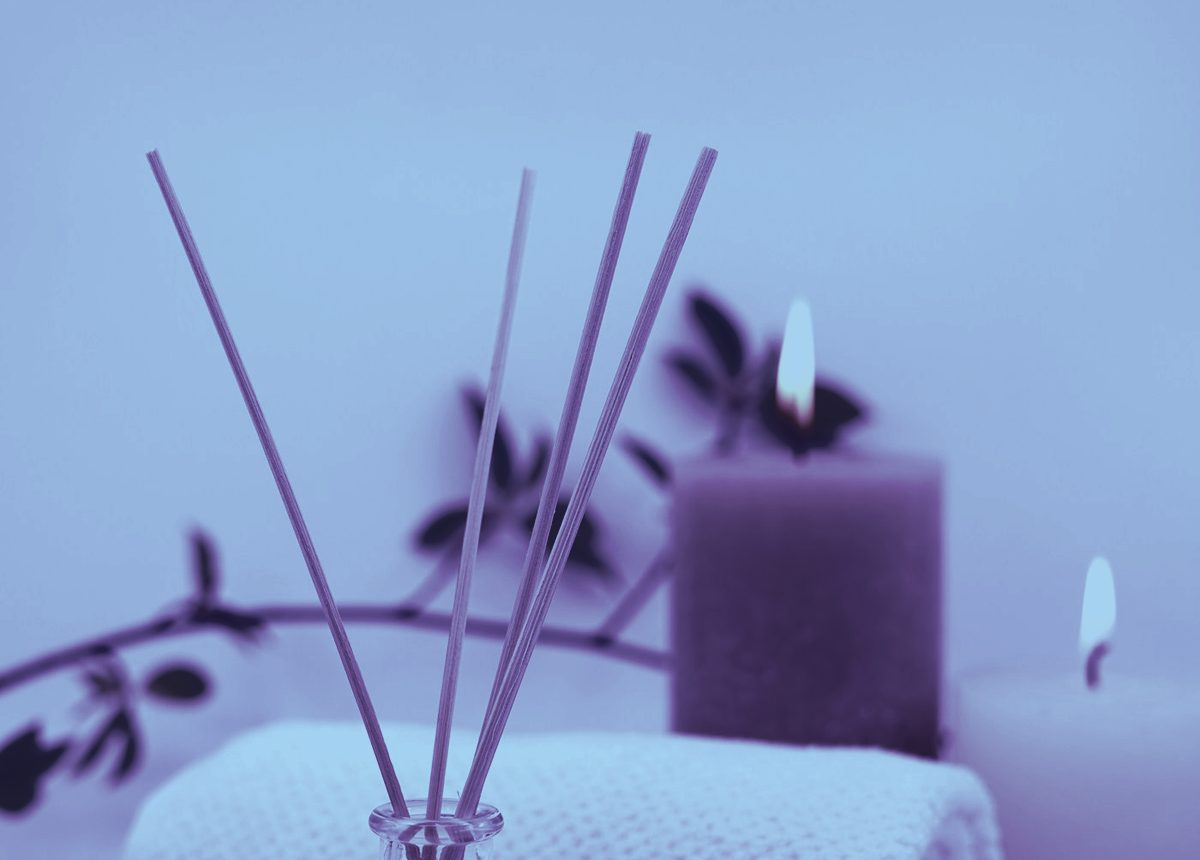 A reed diffuser and softly lit candles on a folded towel, bathed in a calm violet-blue light