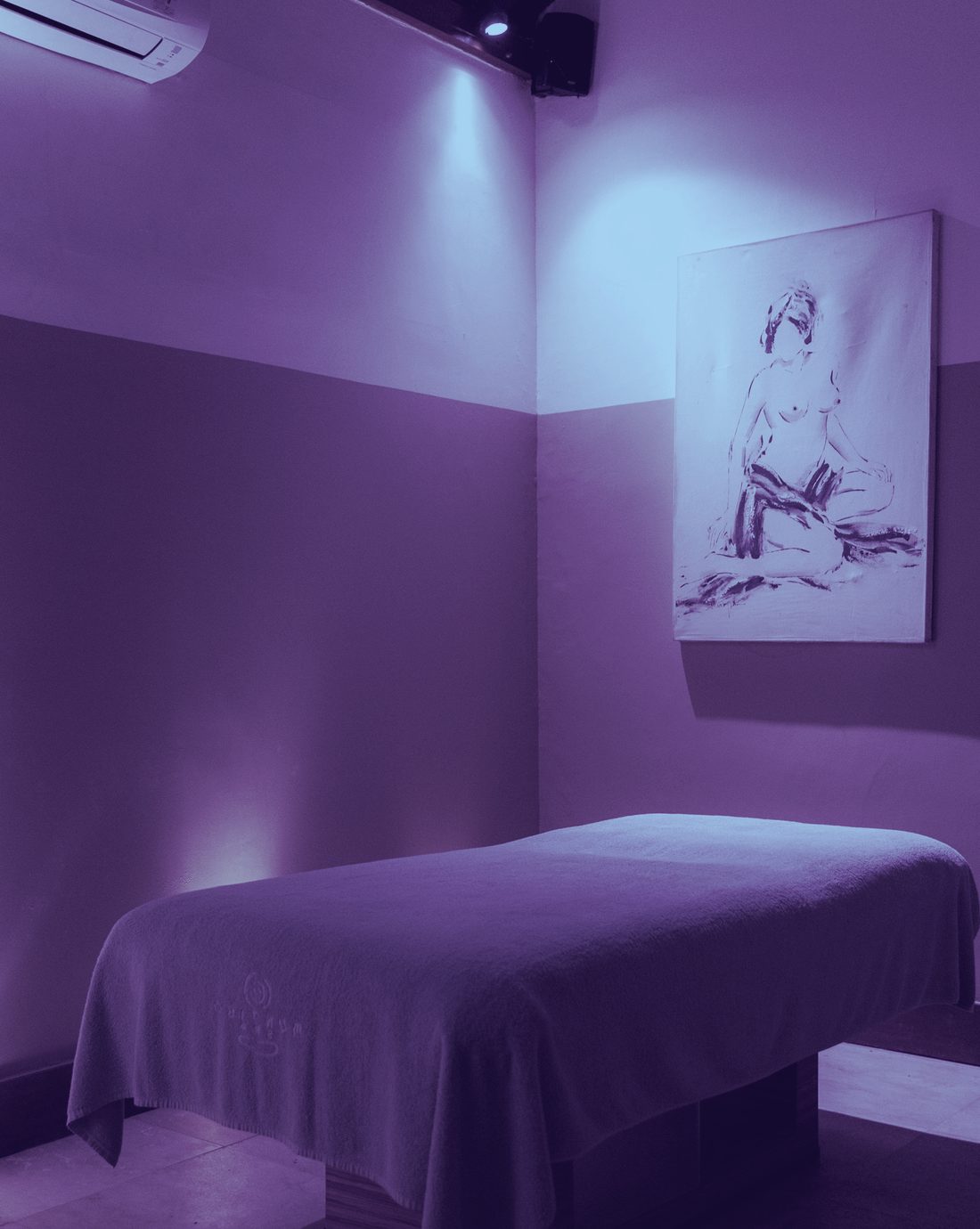 A low-lit, private treatment room with a made massage table, washed in cool violet light